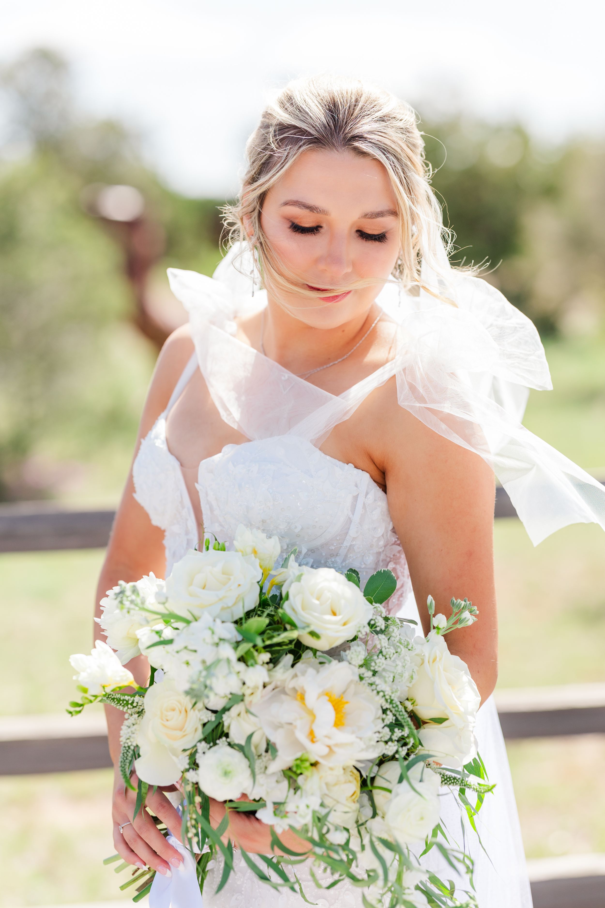 A bride in a white wedding dress holds an elegant bouquet of white roses and greenery on a sunny outdoor day.