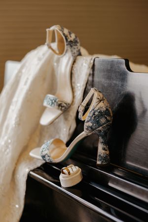 Elegant bridal accessories featuring sparkly silver heels, wedding rings, and white lace dress displayed on piano keys.