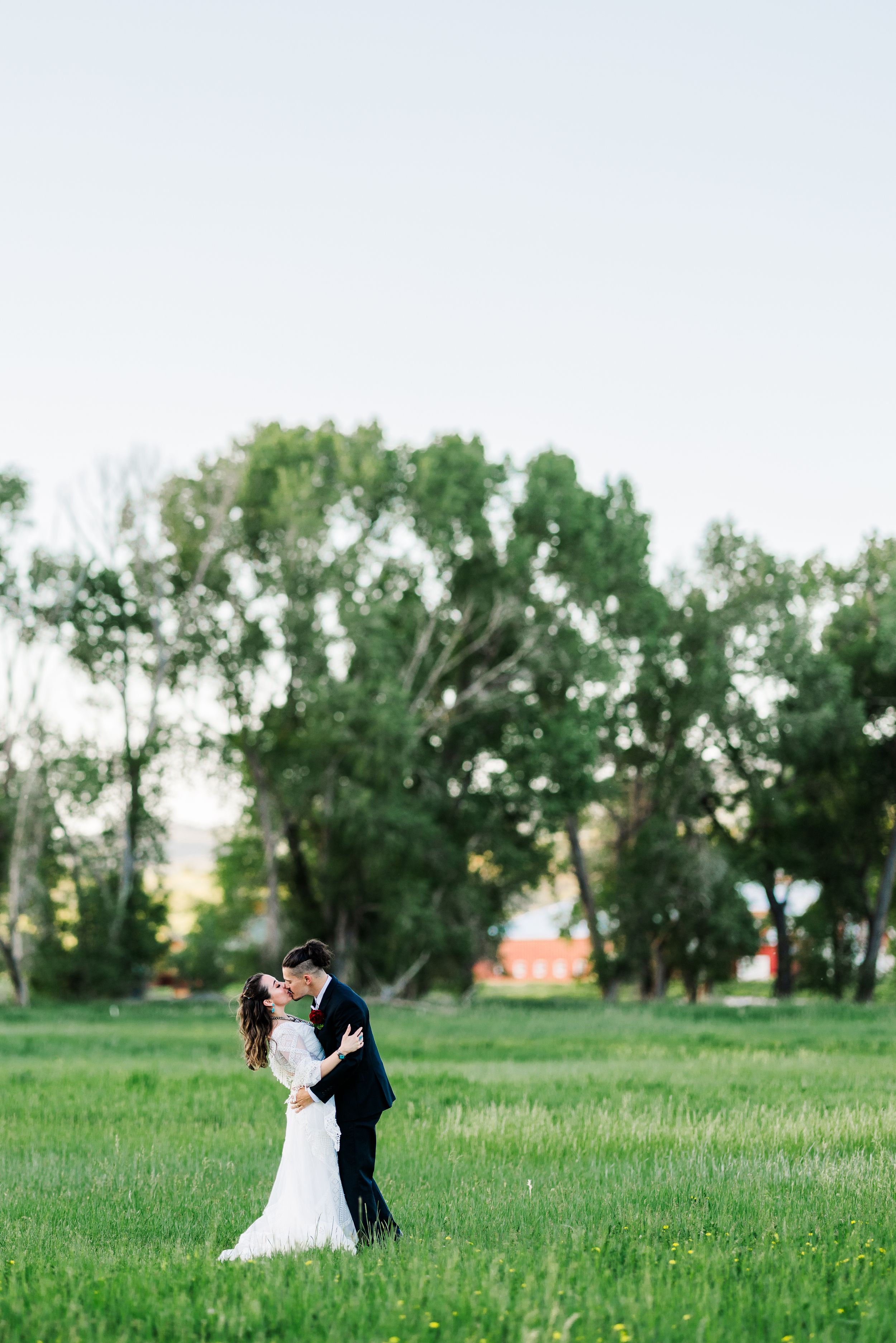 A couple embraces in a green meadow with tall trees in the background during a romantic outdoor photo session.