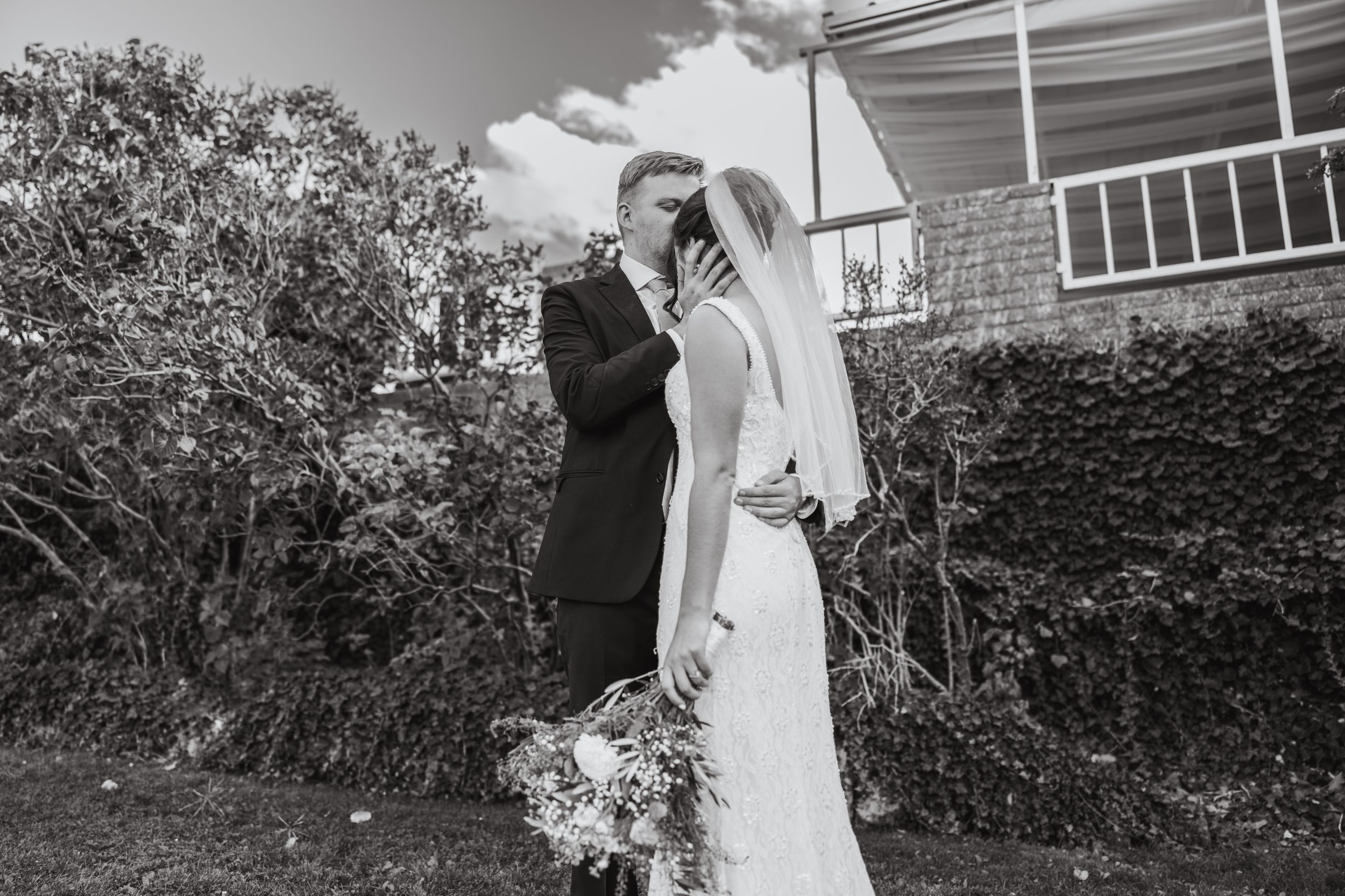 A black and white wedding photograph shows a couple embracing outdoors near a brick building and garden.