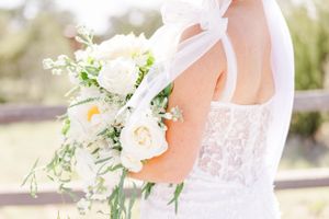 Delicate white bridal bouquet with roses and greenery held against a lace wedding dress in soft sunlight.