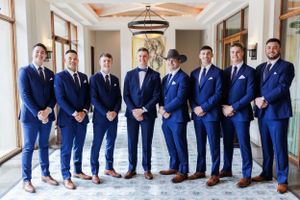 A group of groomsmen in matching navy blue suits standing in a row inside an elegant hallway with a chandelier.