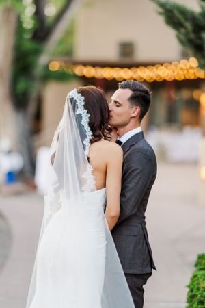 A romantic outdoor wedding scene with a couple kissing against a backdrop of twinkling lights and greenery.