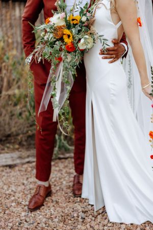 A wedding couple stands together showing burgundy suit and white dress with a vibrant wildflower bouquet with sunflowers.