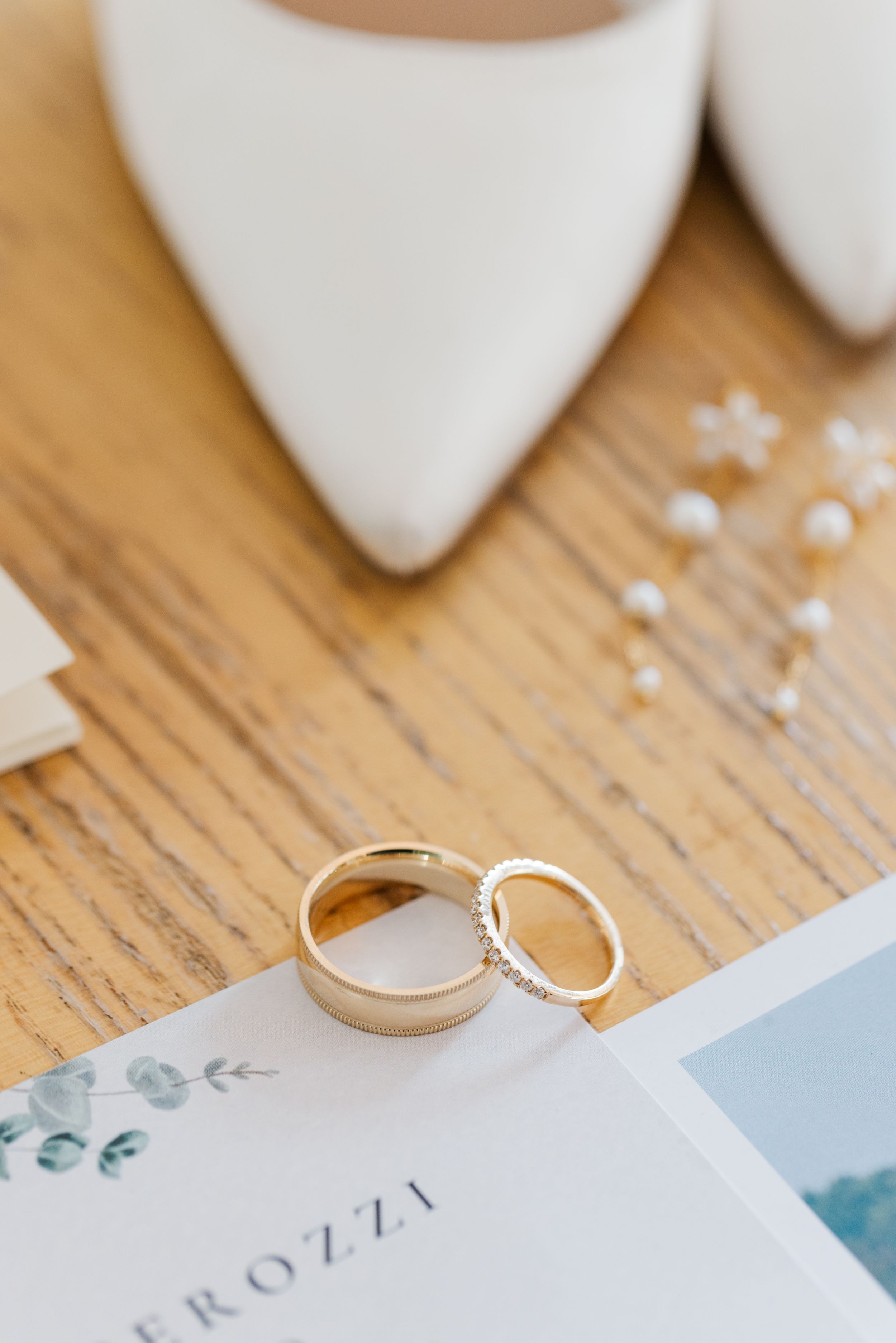 Wedding rings and white bridal shoes arranged with text on a rustic wooden surface with delicate floral accents.