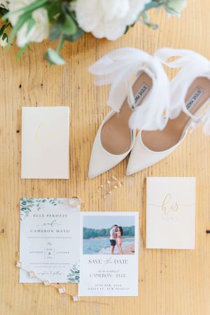 White wedding shoes and invitation cards with greenery accents arranged on a wooden surface for wedding photography.