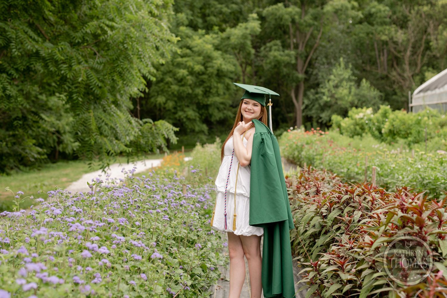 Graduate in green academic regalia poses along garden path with purple wildflowers.