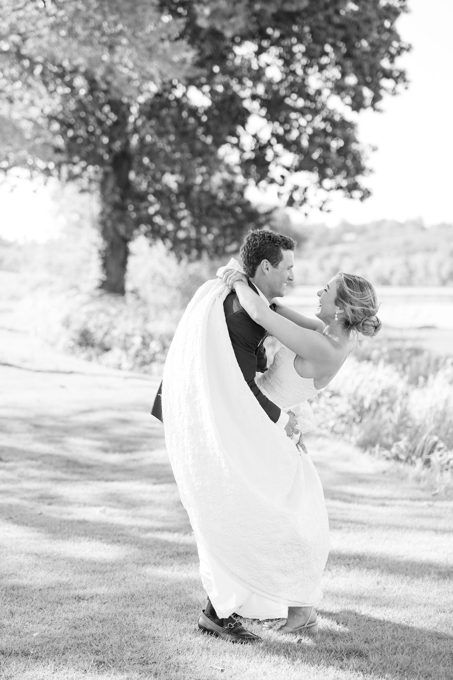A bride and groom share a romantic dance outdoors under a tree in black and white photography.