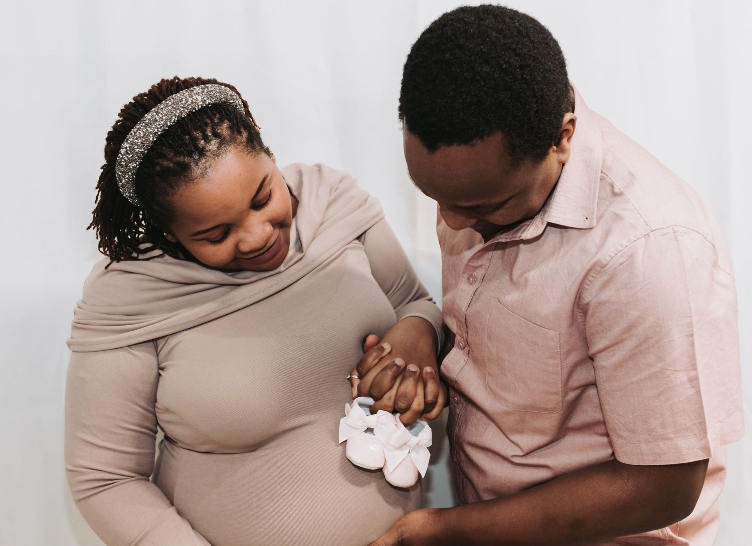 Expecting couple holds small baby shoes while sharing a tender moment against white background.