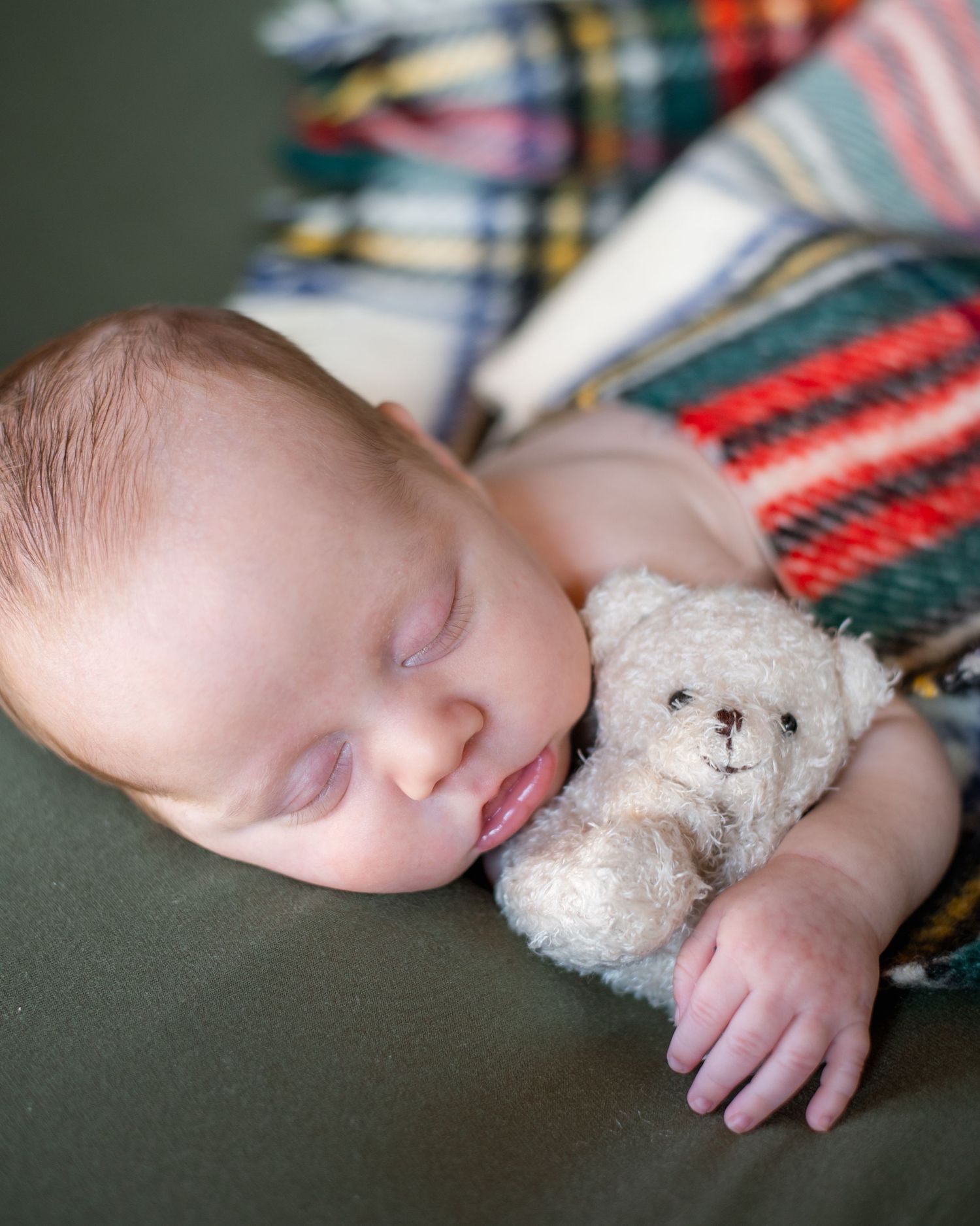 Sleeping infant cuddled up with a small white teddy bear on a couch with colorful plaid blankets.