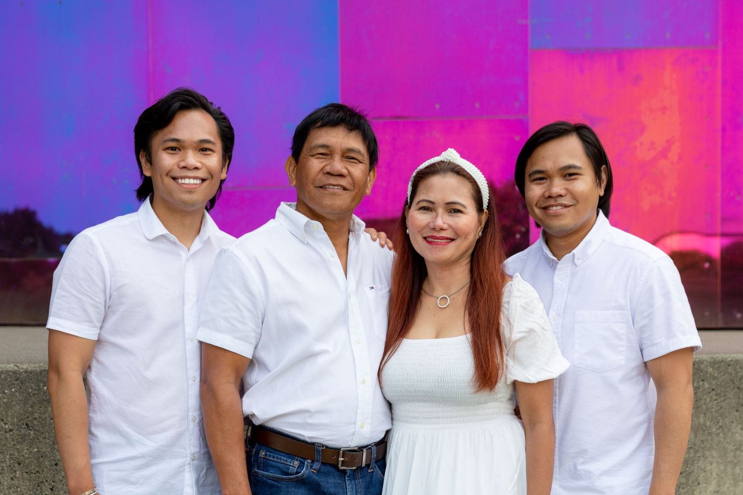 A group wearing coordinated white outfits poses together against a vibrant purple and pink backdrop.