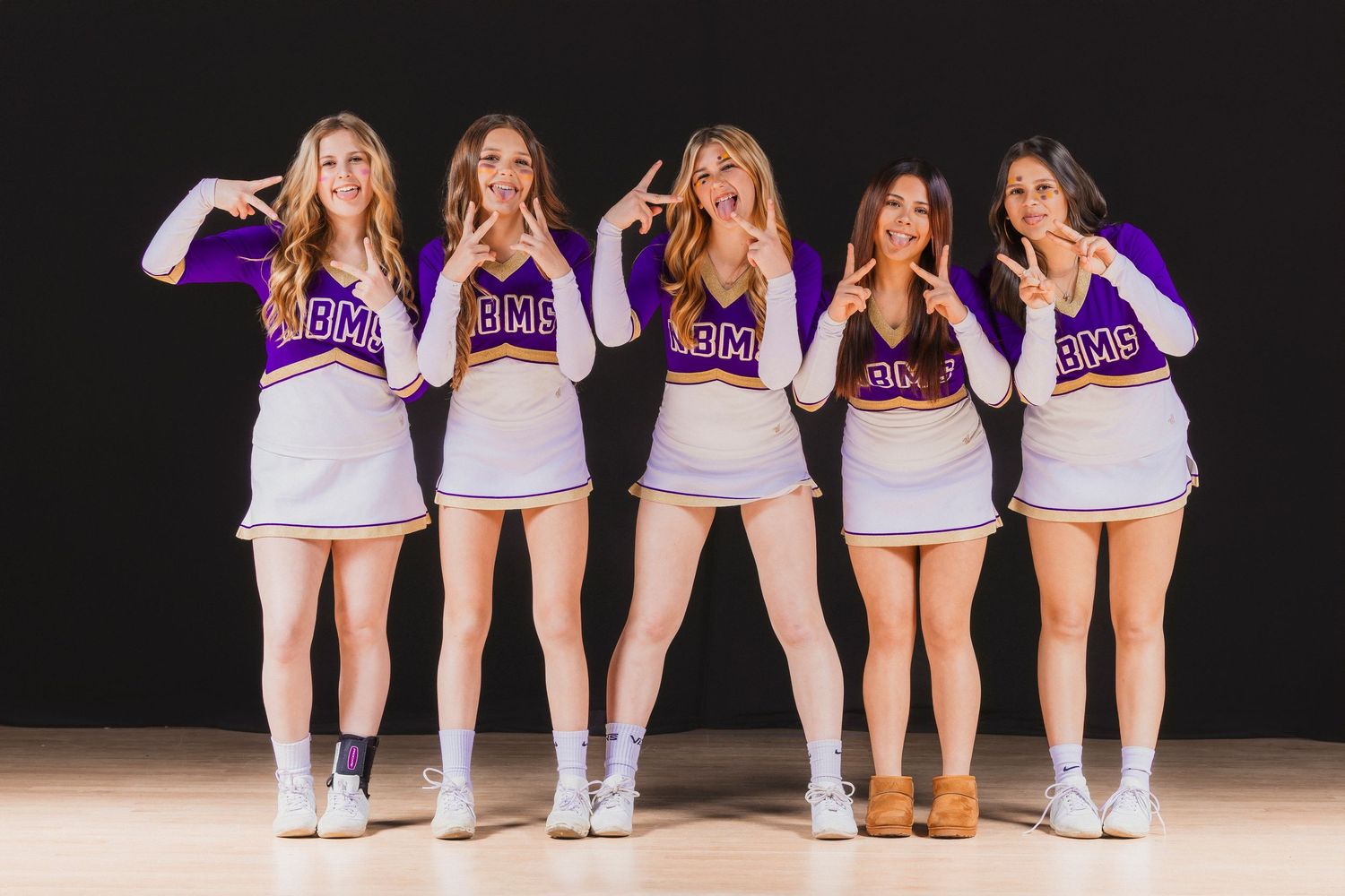 Group of cheerleaders in purple and white uniforms pose together on dark stage.