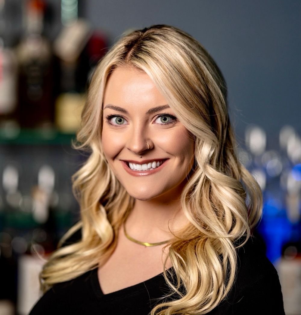 A professional headshot of a person with wavy blonde hair wearing a black top, smiling warmly against a dark background.
