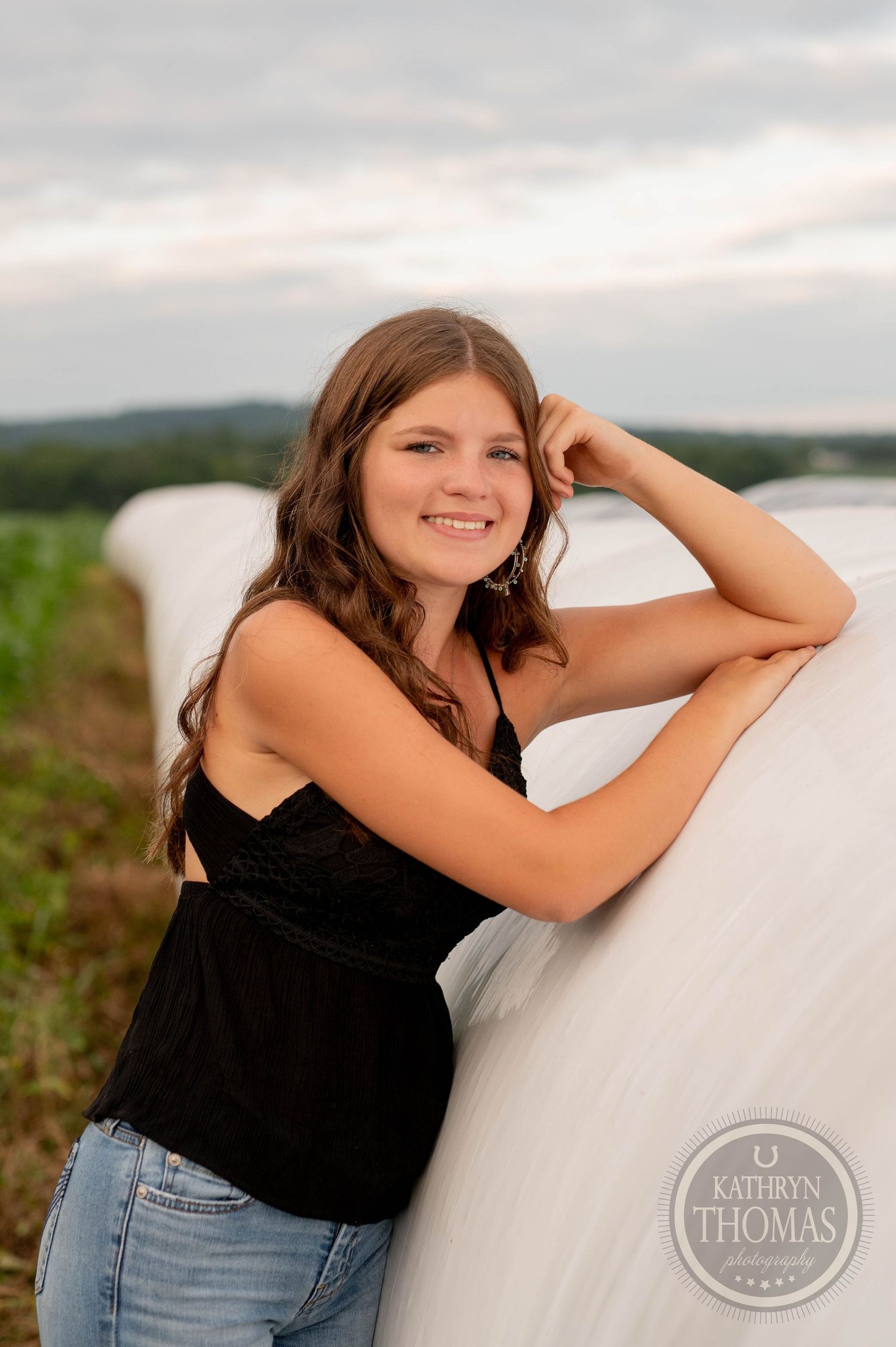 A casual portrait against round bales wearing jeans and black tank top in evening light.