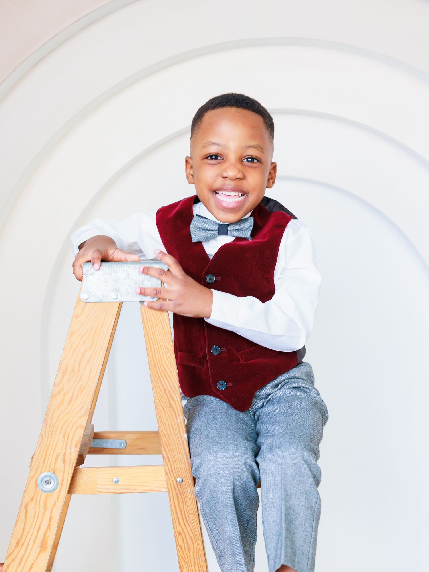 Smiling boy in a white shirt, red vest, and bow tie sitting on a wooden ladder with a curved white backdrop. Toronto family photography