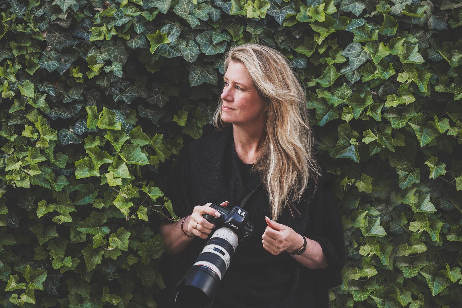 Blonde woman in black outfit holding a DSLR camera with telephoto lens, standing in front of lush green ivy wall.