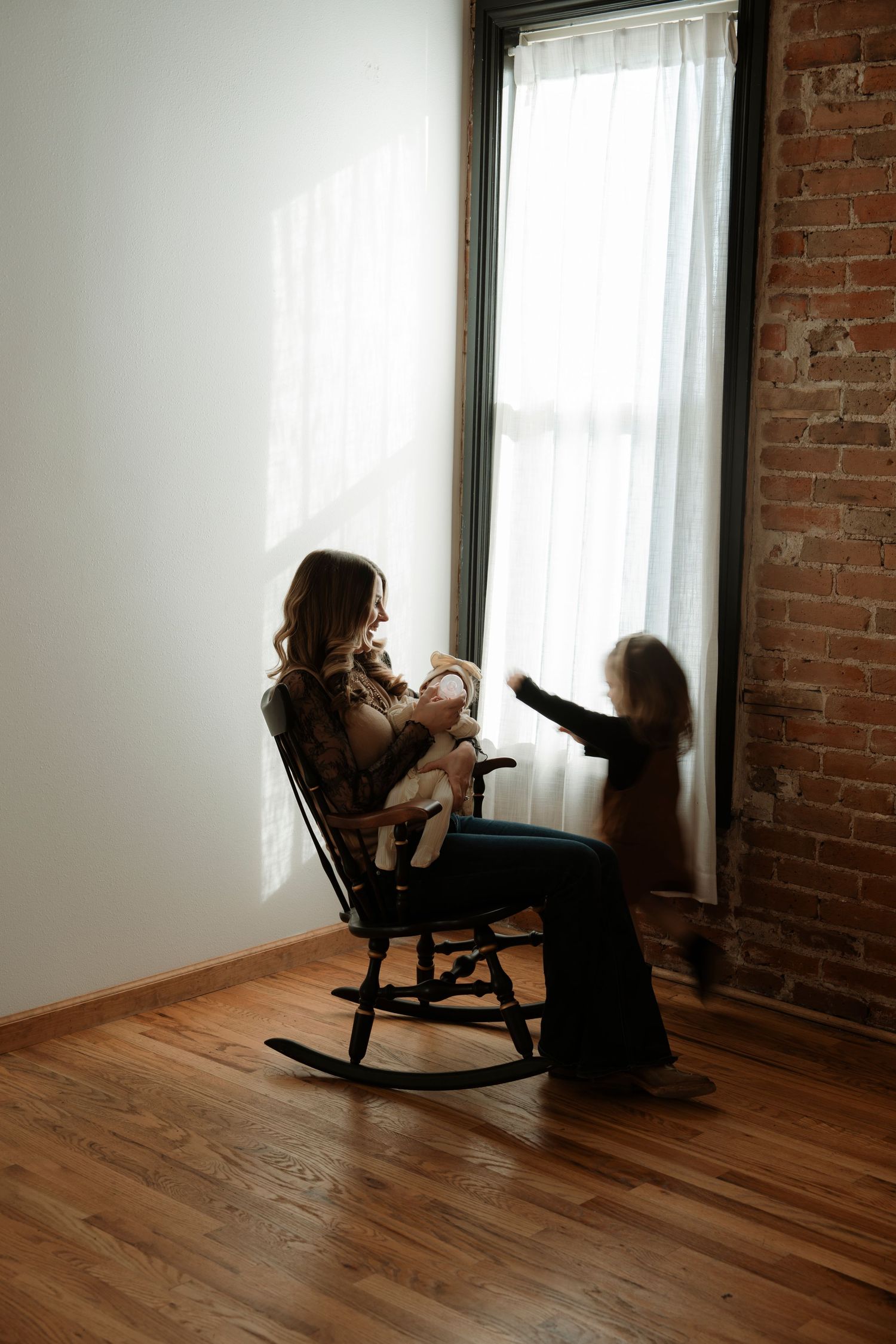 A mother sits in a rocking chair nursing her baby near a sunlit window while a toddler plays with the curtain.