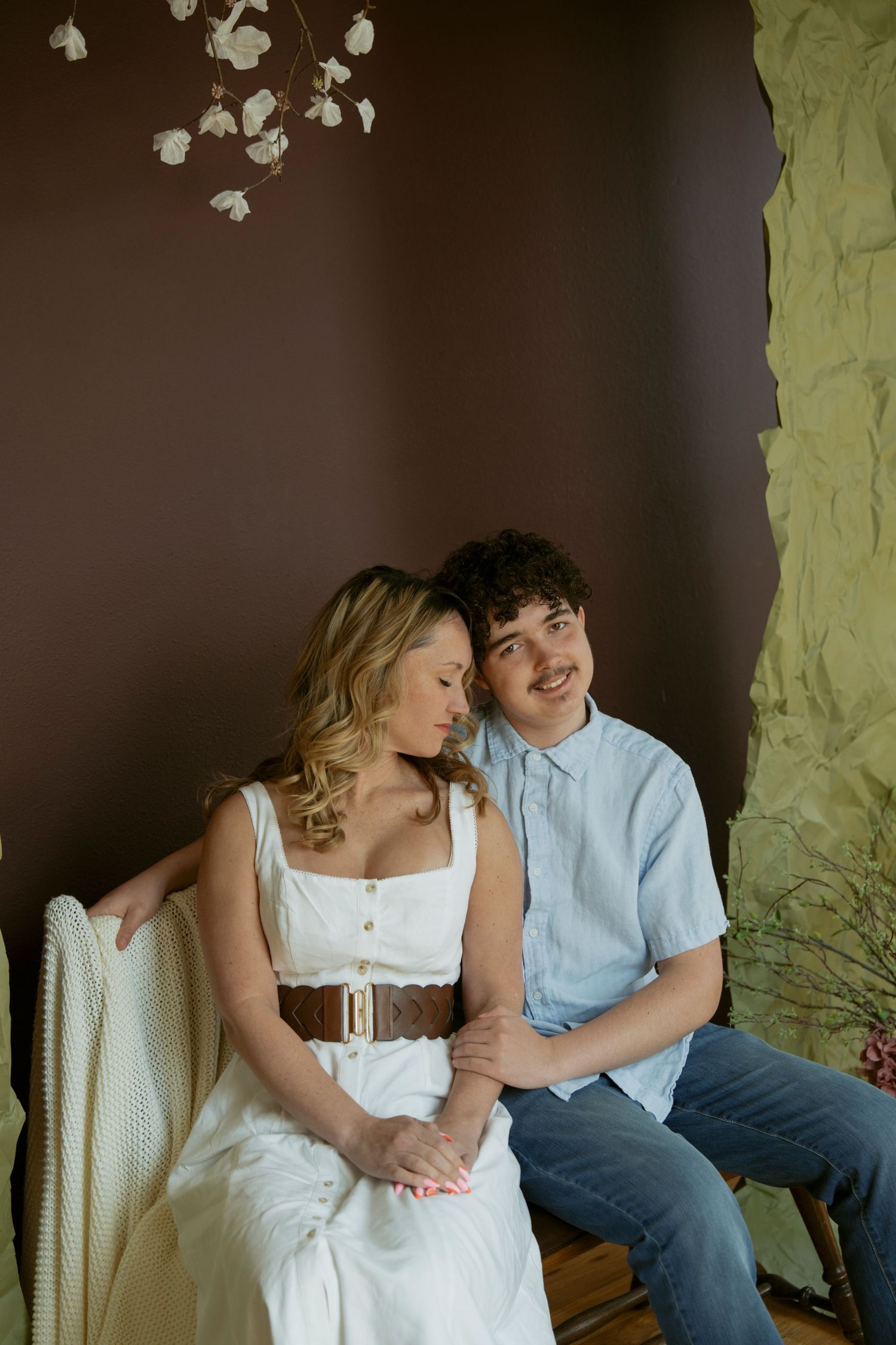 A smiling young couple sits together on a bench, woman in white dress with brown belt, man in light blue shirt and jeans.