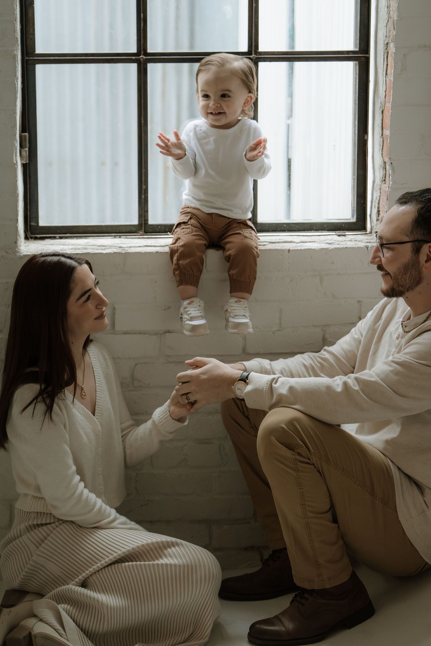 Happy toddler sitting on windowsill with smiling parents on floor in bright white brick room.