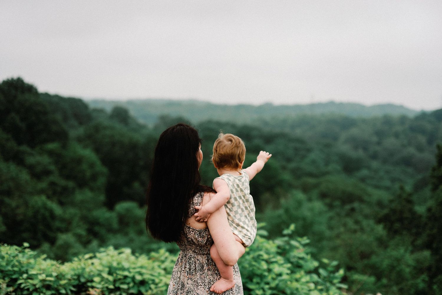 A mother holds her baby while overlooking a lush green forest landscape on a cloudy day.