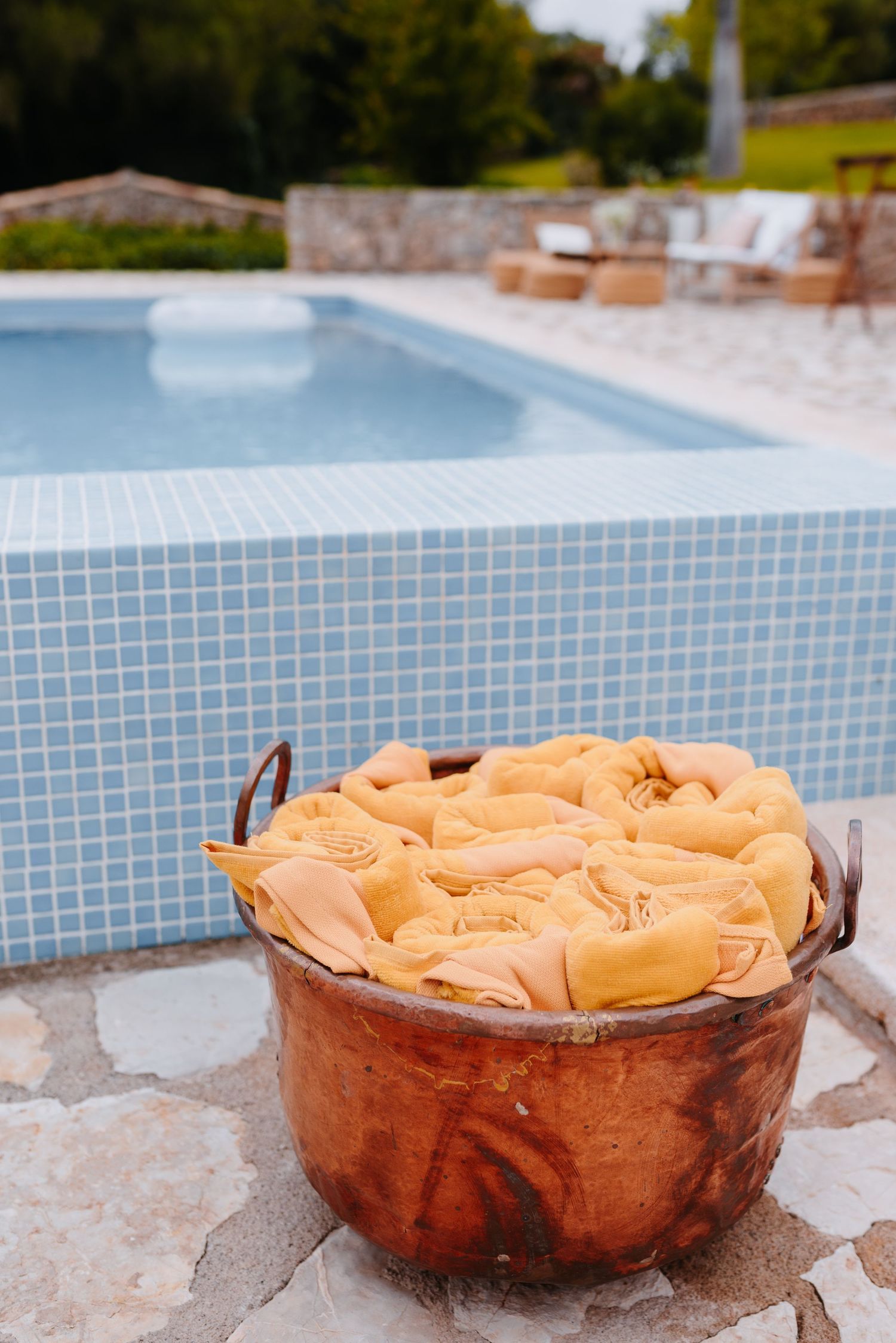 Rustic copper pot filled with folded yellow napkins beside a blue mosaic tile swimming pool in a garden setting.
