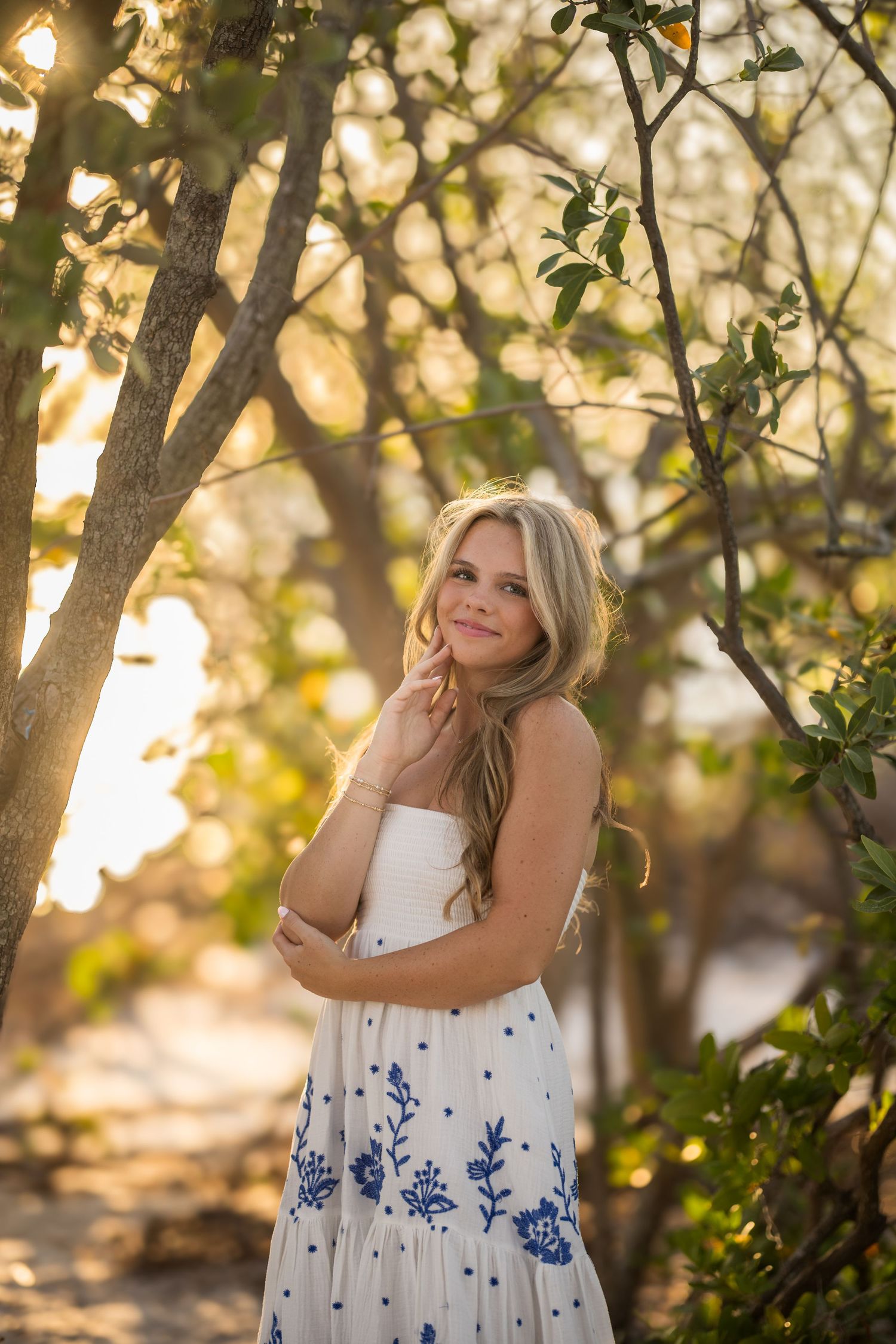 Smiling blonde woman in white floral sundress poses among golden-lit trees at sunset in nature.