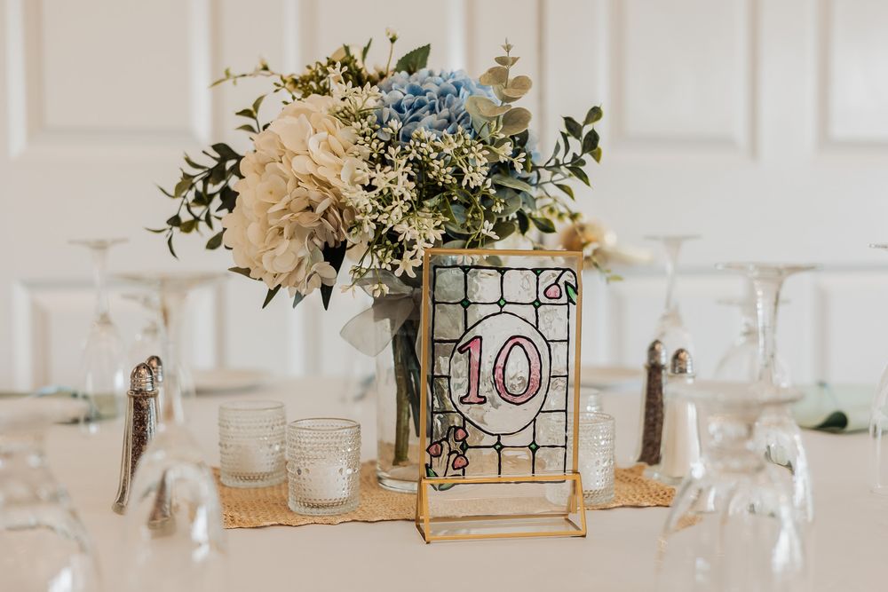 Wedding table centerpiece with white and blue flowers, table number 10 sign, wine glasses, and candles on white tablecloth.