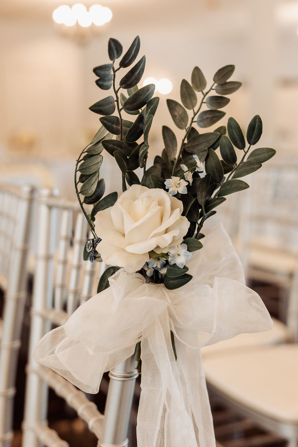 Elegant wedding chair decoration with white rose, greenery, and sheer bow in a softly lit reception venue.
