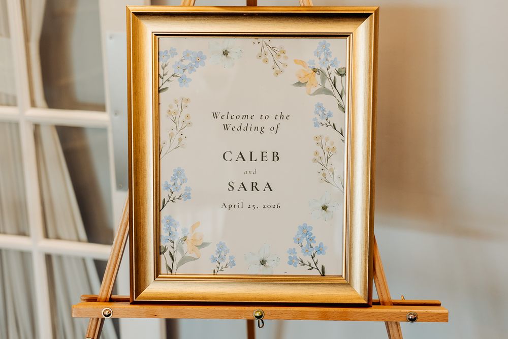 Gold framed wedding welcome sign for Caleb and Sara on a wooden easel with blue floral accents.