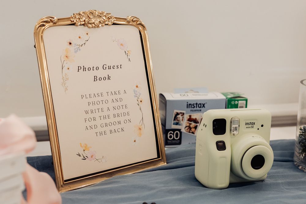 Gold framed Photo Guest Book sign beside a mint Fujifilm Instax Mini camera and film at a wedding reception.
