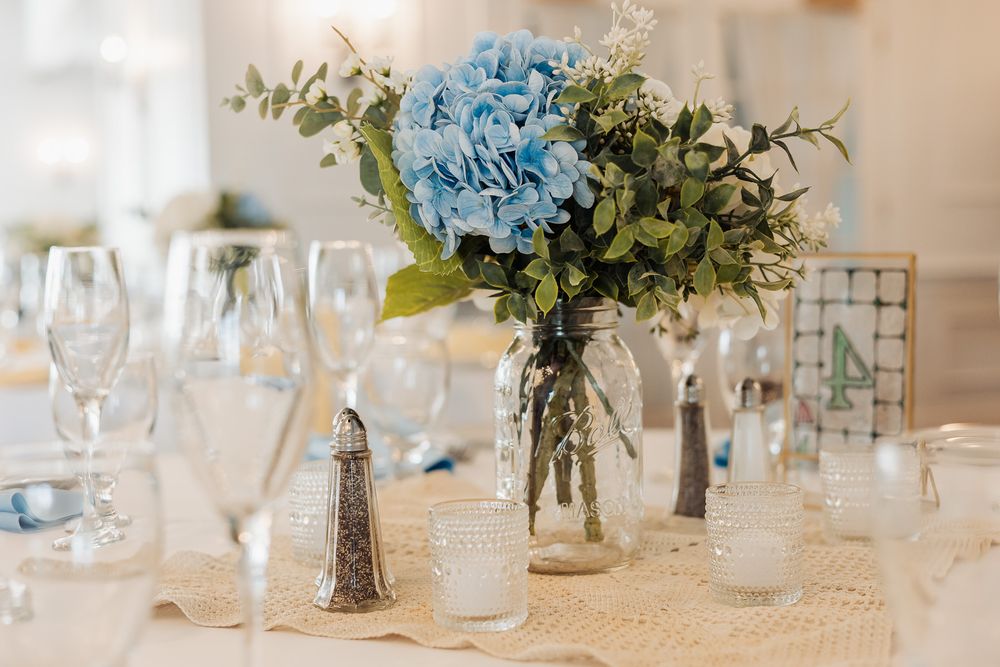 Blue hydrangea centerpiece in mason jar on burlap table runner with elegant glassware at wedding reception.
