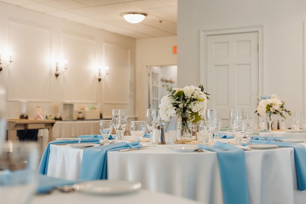 Elegant wedding reception tables with blue napkins, white linens, floral centerpieces, and crystal glassware in a bright venue.
