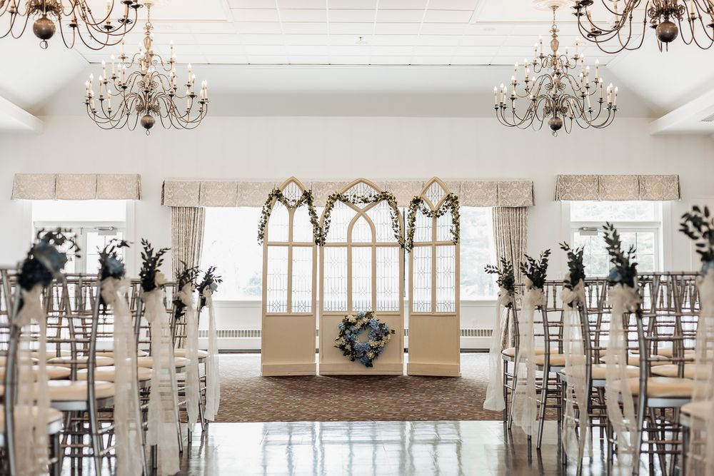 Elegant wedding ceremony venue with chandelier lighting, chiavari chairs, floral aisle decor, and a wooden arch backdrop.
