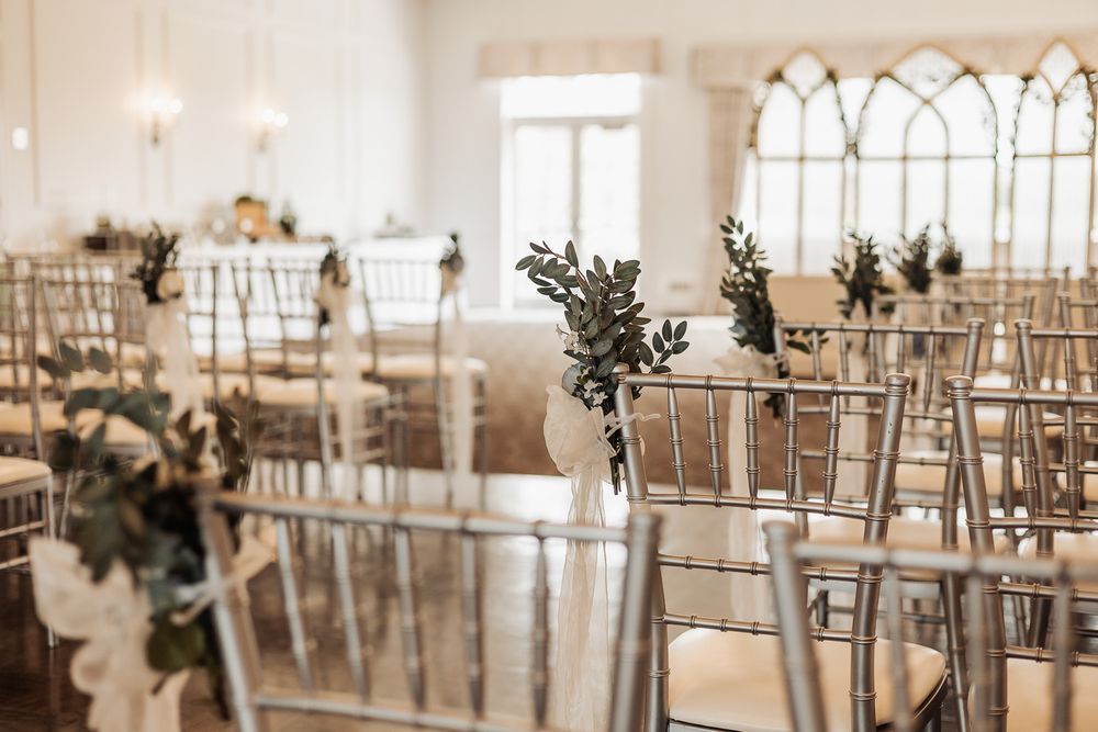 Elegant wedding ceremony setup with silver chiavari chairs adorned with greenery in a bright white venue.