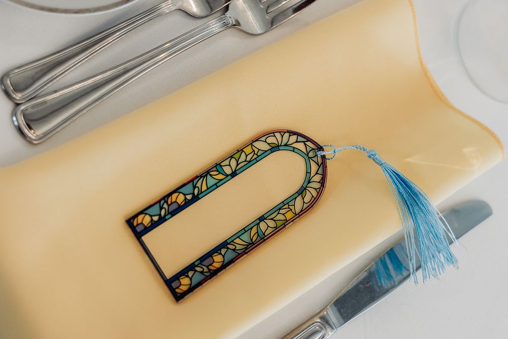 Decorative bookmark with colorful stained glass pattern and blue tassel resting on a cream plate with silverware.