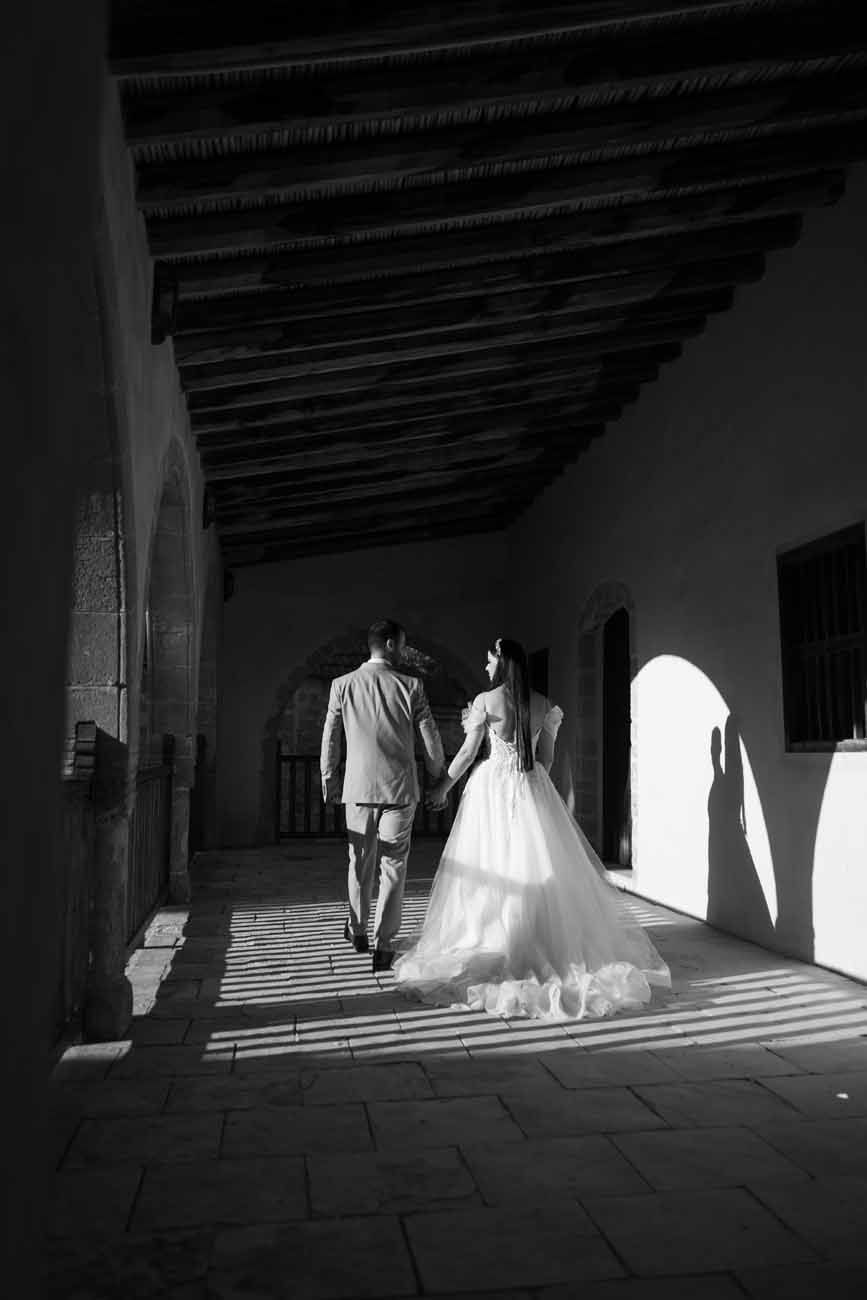 Black and white photo of a bride and groom walking hand in hand through a sunlit colonial corridor.