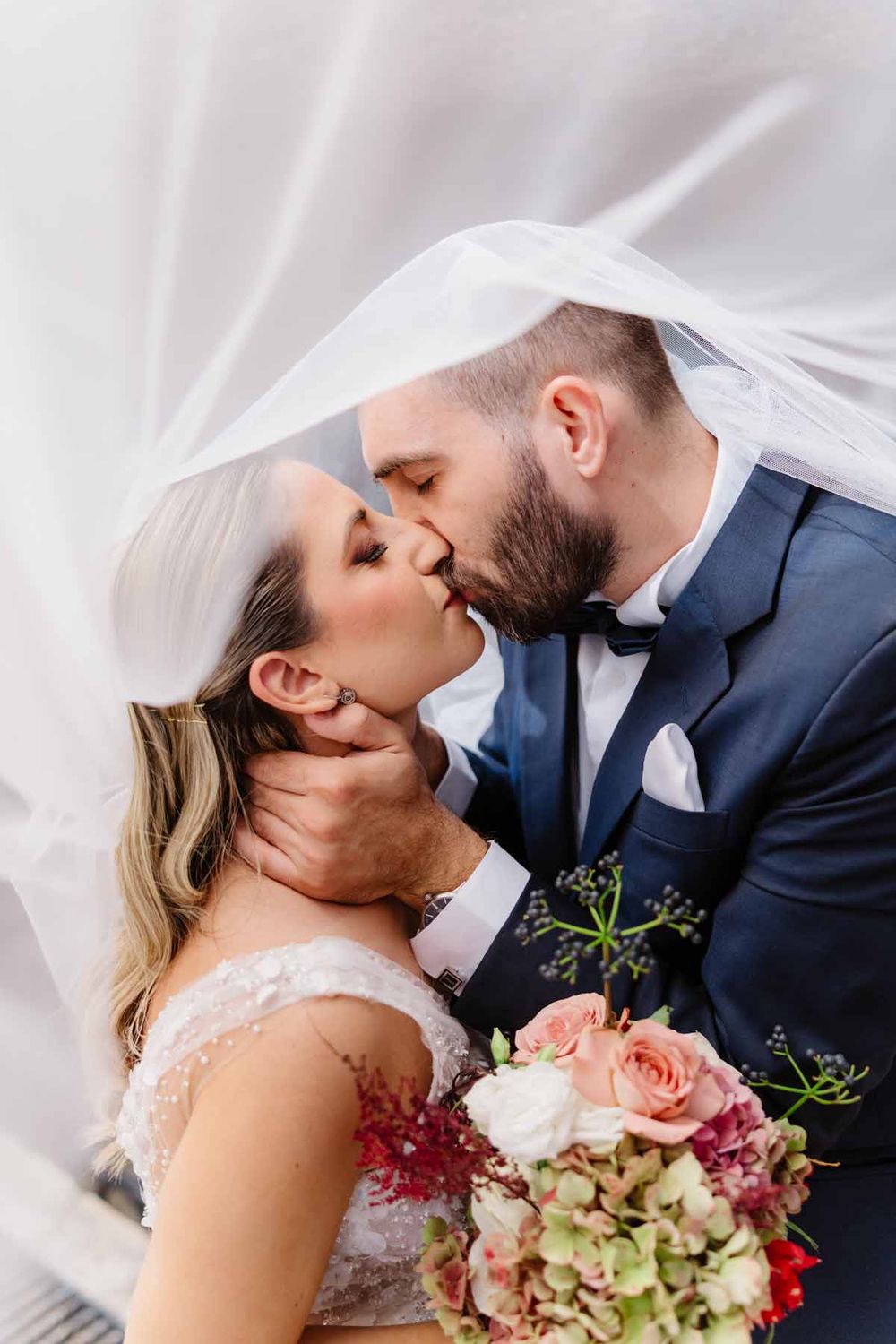 Bride and groom sharing a romantic kiss under a flowing white veil, bride holding a pink and green floral bouquet.