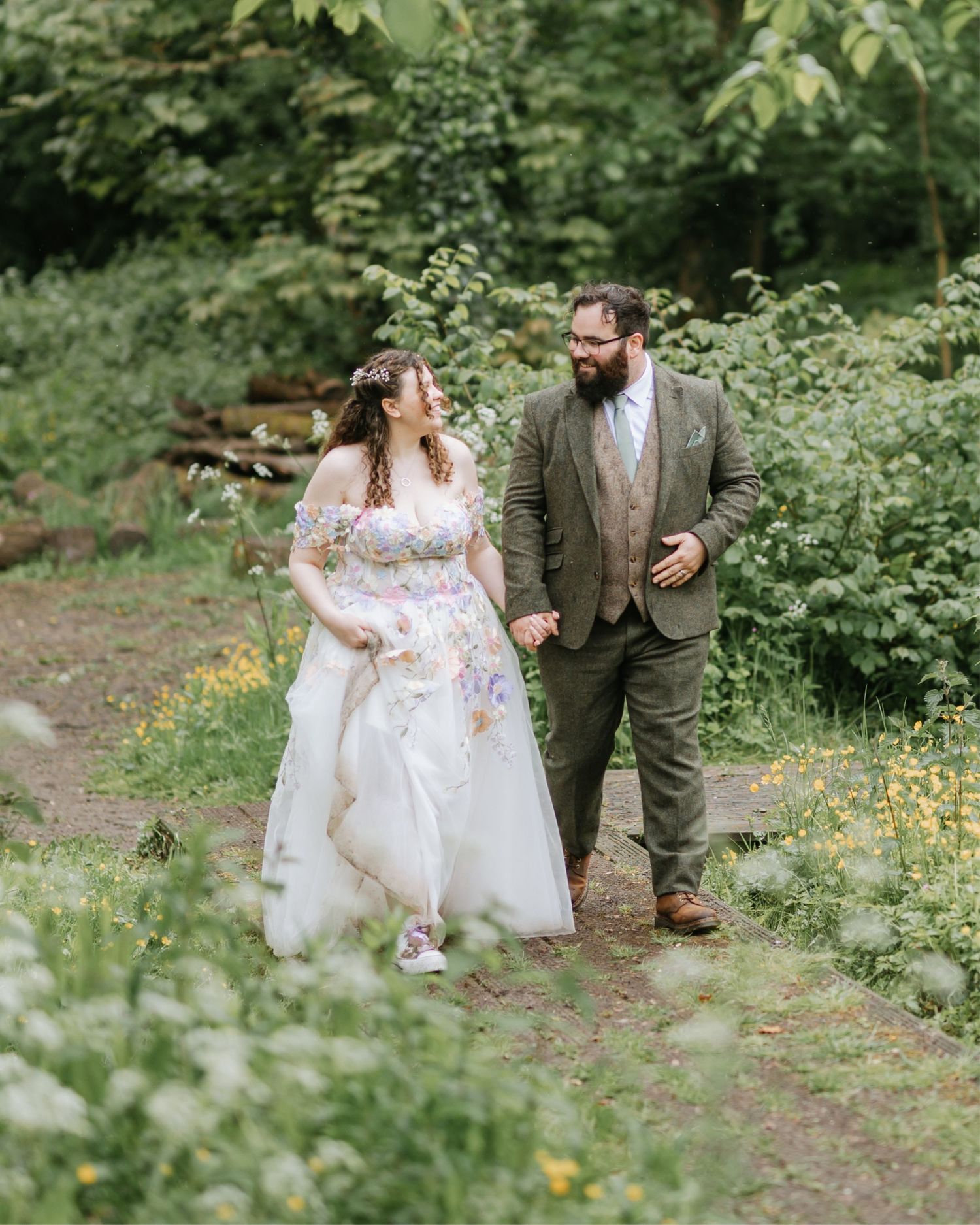 Bride and groom walk hand-in-hand through lush green woodland, bride wearing floral wedding dress, groom in olive suit.
