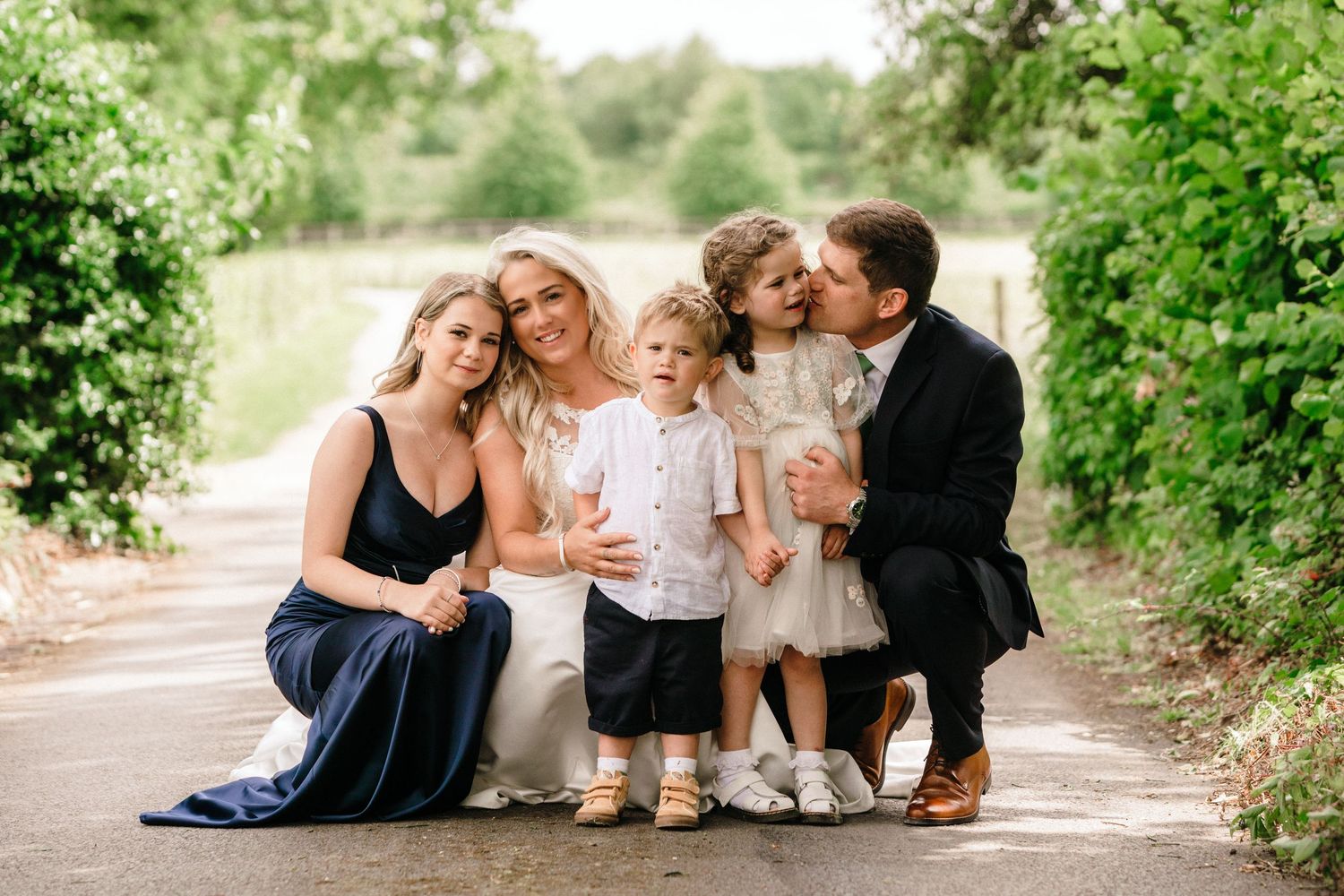 Family portrait with bride, groom, two young children, and bridesmaid posing on a tree-lined path.