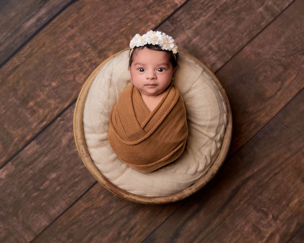 Newborn baby swaddled in a warm brown wrap, wearing a floral headband, posed in a wooden bowl on a rustic floor.