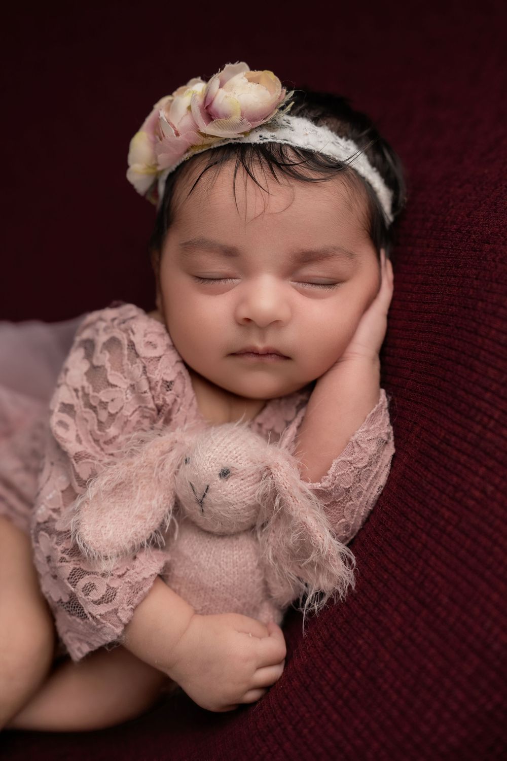 Sleeping newborn baby girl in pink lace outfit holding stuffed bunny with floral headband on dark red background.