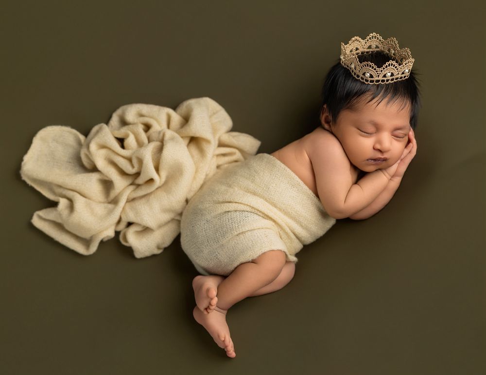 Sleeping newborn baby wearing a gold crown, wrapped in cream fabric with a floral backdrop on olive green.