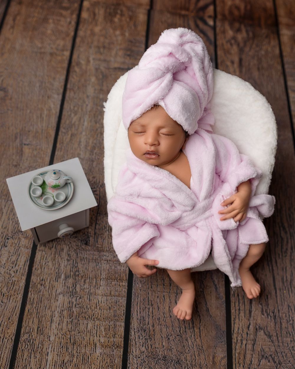 Sleeping newborn baby wrapped in pink fluffy robe and towel turban, posed in tiny white chair beside miniature spa table.