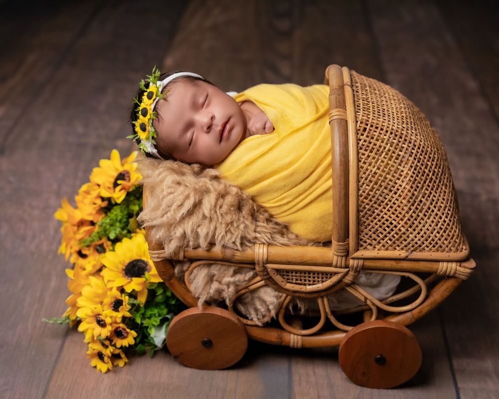 Newborn baby wrapped in yellow swaddle sleeping in wicker pram with sunflowers and floral headband.