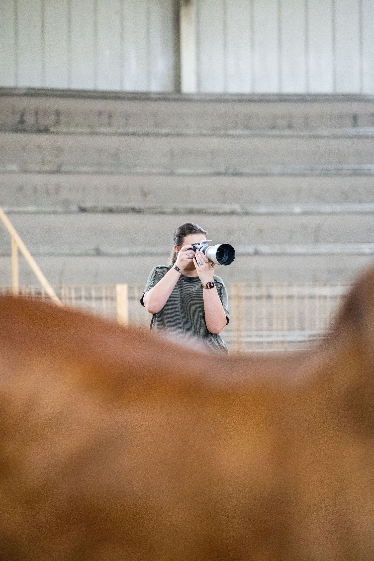 Photographer with telephoto lens capturing photos inside a barn with a brown horse in the foreground.