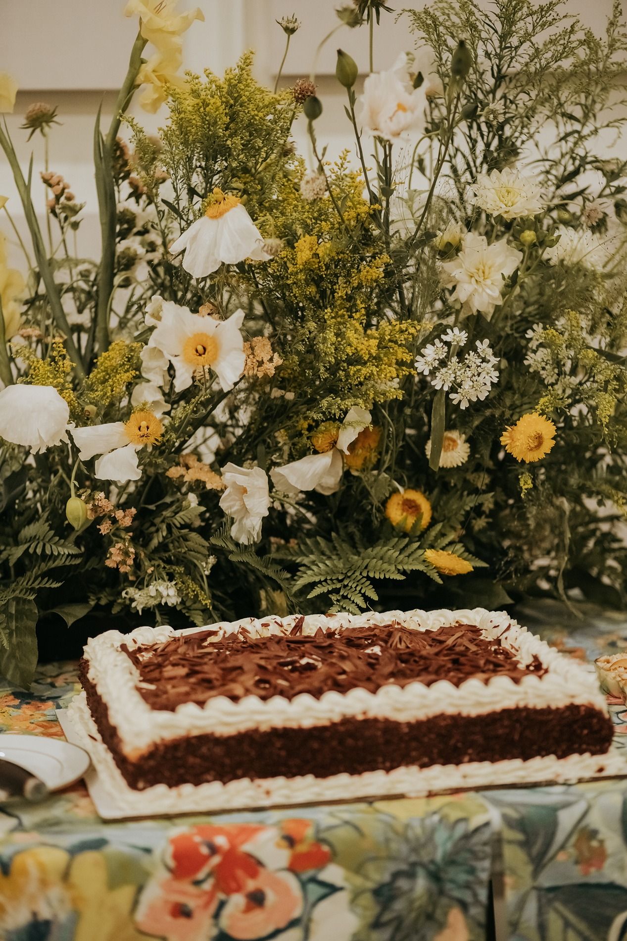 Rectangular chocolate cake with white frosting displayed on a floral tablecloth beside a lush bouquet of white and yellow flowers.