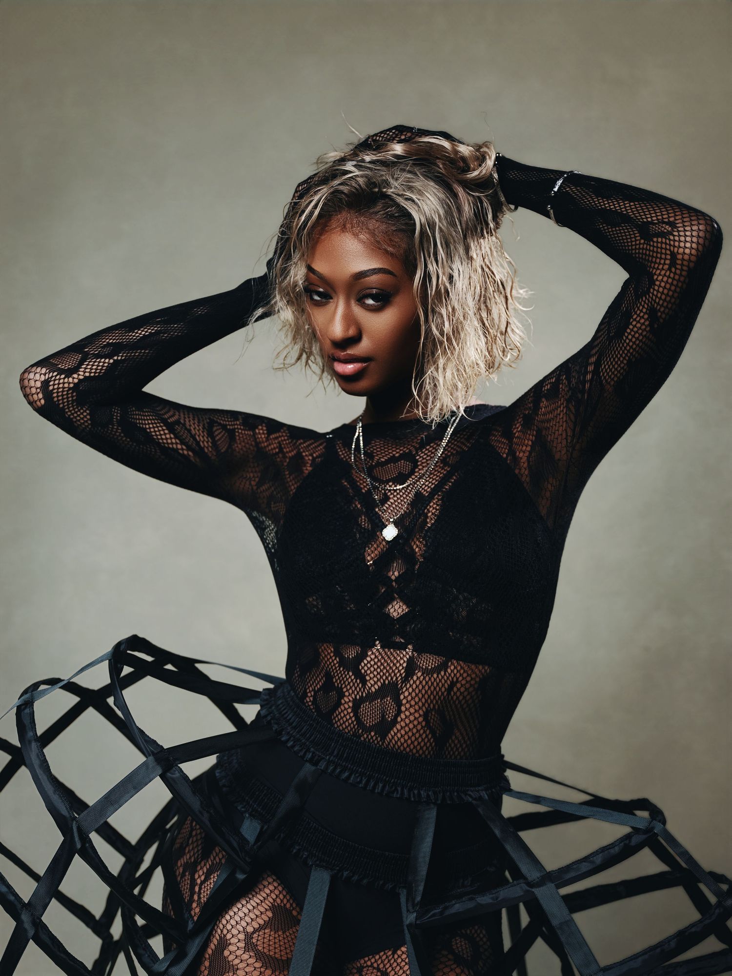 A woman in a black lace bodysuit and structured hoop skirt poses confidently with hands behind her head.