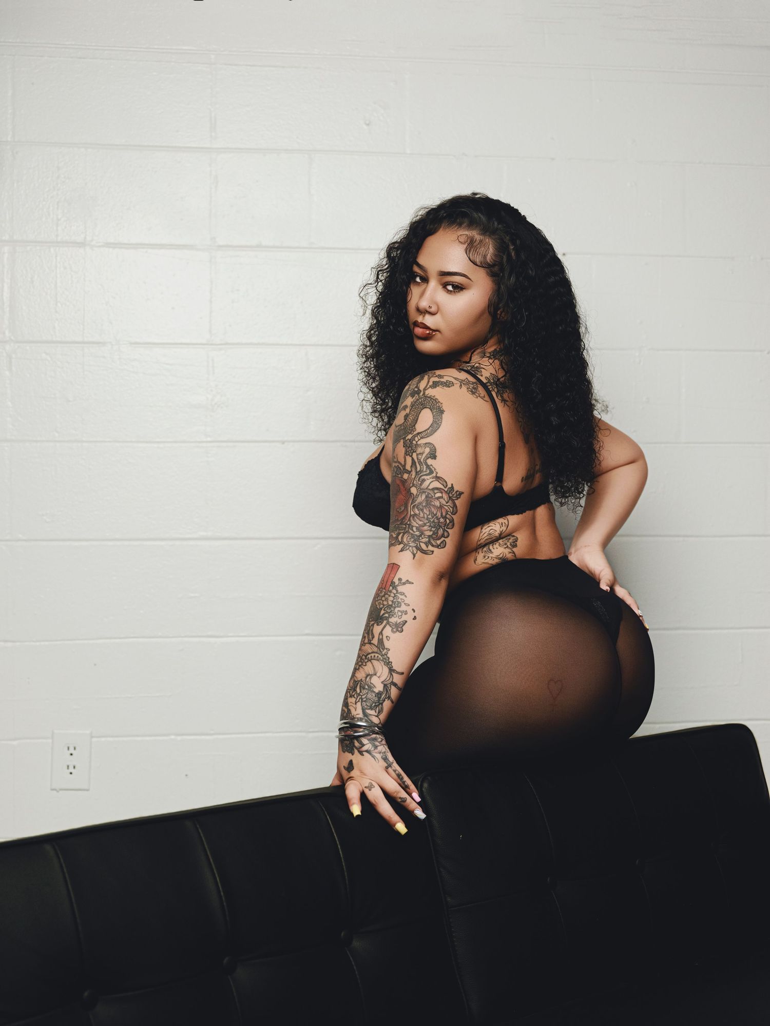 Tattooed woman with curly hair posing in black lingerie and sheer leggings against a white brick wall.