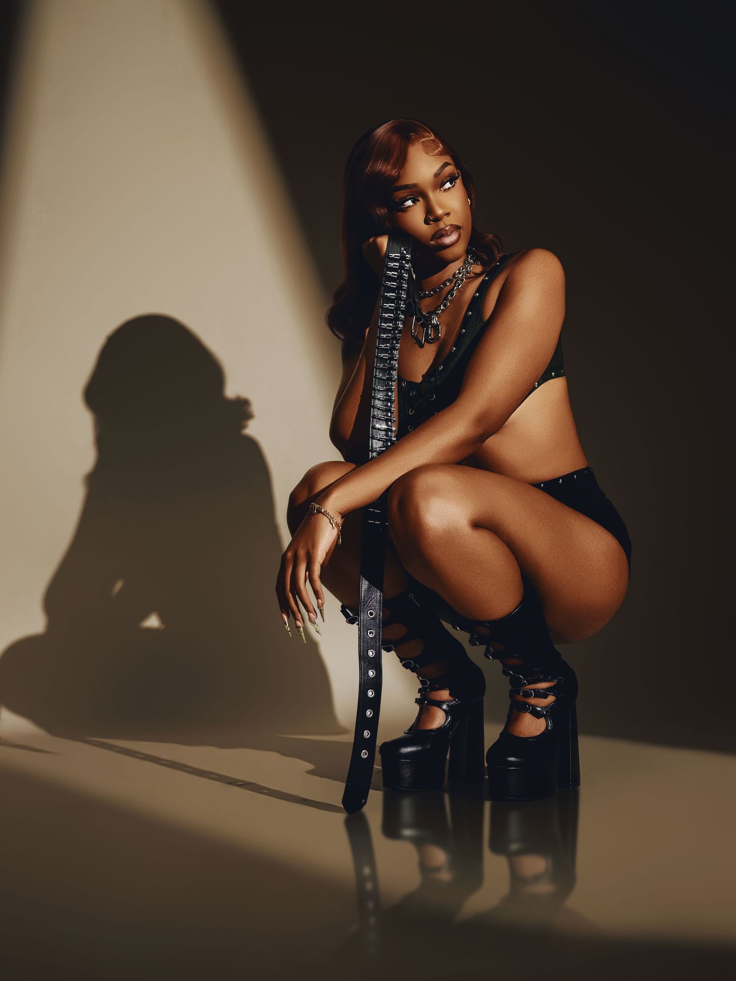 Woman in black lingerie and platform boots crouches dramatically in studio lighting with shadow on wall.