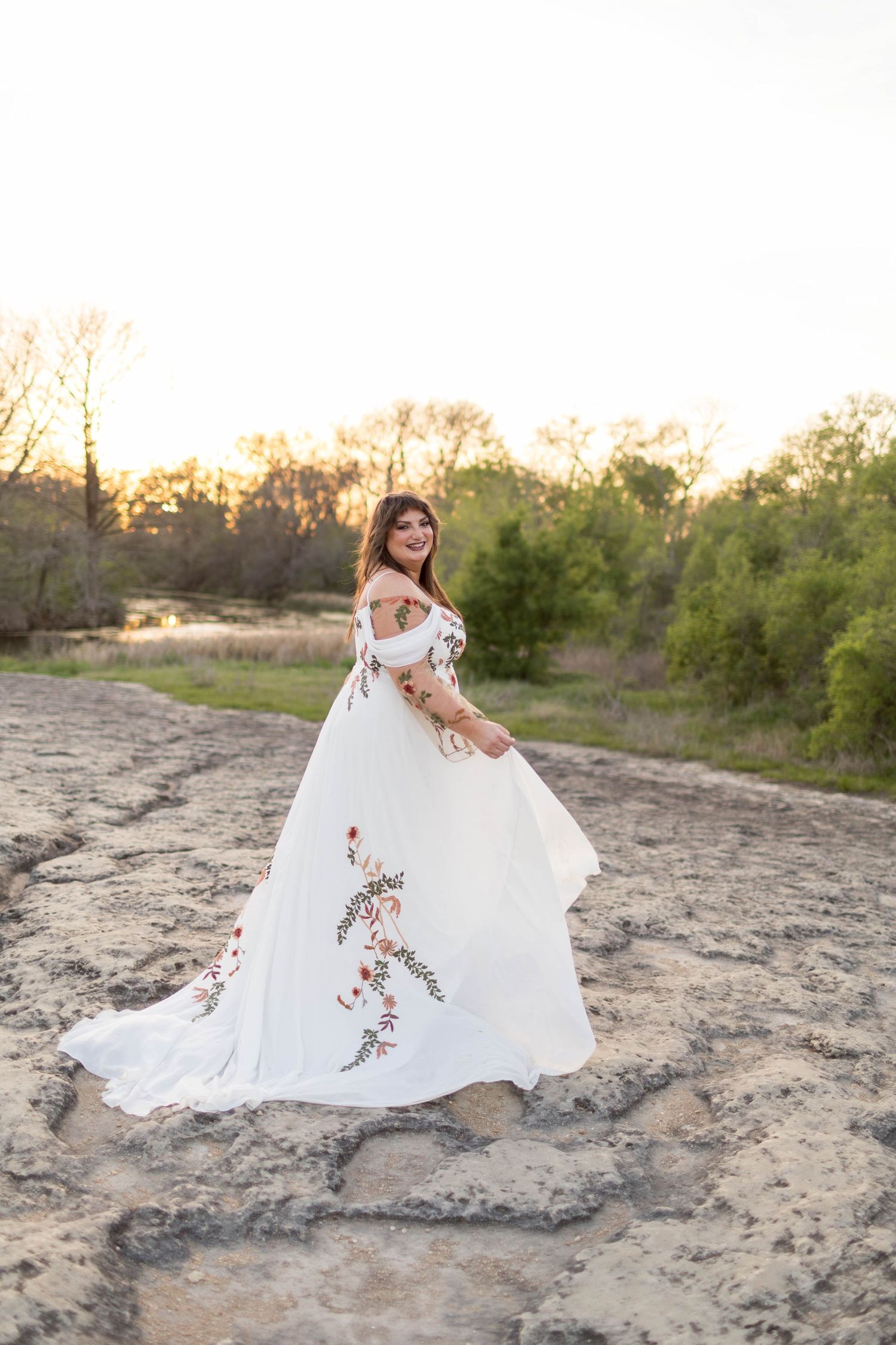 Bride in white floral embroidered off-shoulder gown twirling outdoors at sunset with trees in background at McKinney Falls.